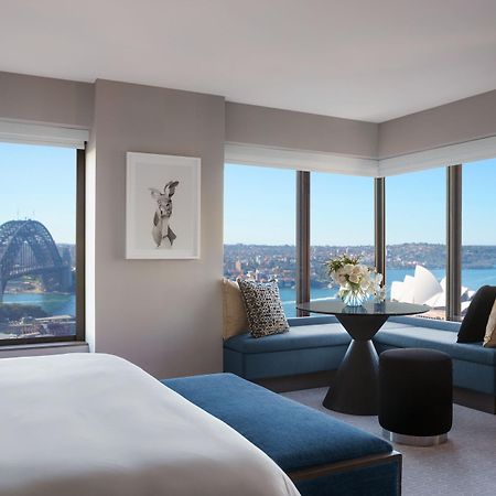 Four Seasons Hotel Sydney Exterior photo The photo shows a modern hotel room with large windows offering a panoramic view of a city skyline. In the foreground, there is a bed with white linens and a blue accent bench. A piece of artwork is displayed on the wall. In the corner, there is a ci