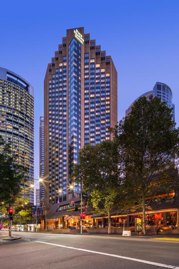 Four Seasons Hotel Sydney Exterior photo The photo shows a tall modern building with a glass façade, likely an office or residential space, illuminated in the evening. In the foreground, there are trees and streetlights, contributing to a lively urban atmosphere. Surrounding the main buildi