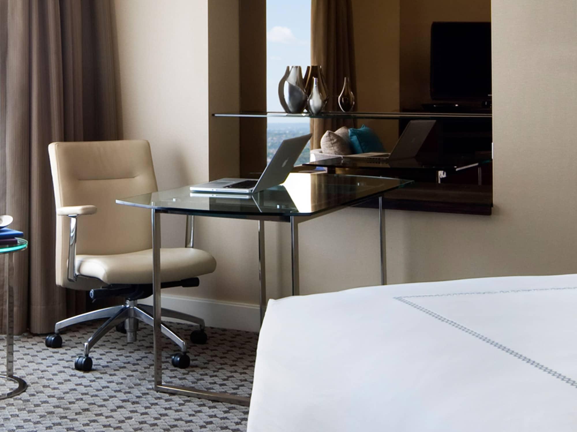 Four Seasons Hotel Sydney Exterior photo The photo depicts a modern hotel room setup. On one side, there's a sleek glass desk with a laptop set up on it, accompanied by a comfortable, ergonomic chair. The desk is positioned next to a window draped with curtains, and there are decorative ite