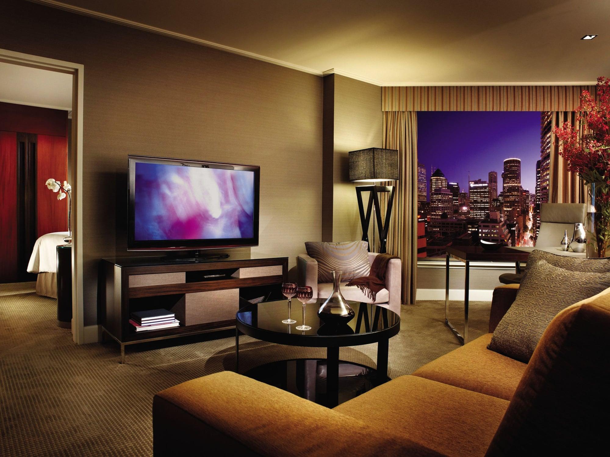 Four Seasons Hotel Sydney Room photo The photo showcases a stylish hotel room or a modern living area. It features a comfortable brown sofa with orange cushions, a glass coffee table, and a sleek television mounted on a media console. The room is well-lit, and large windows provide a vi