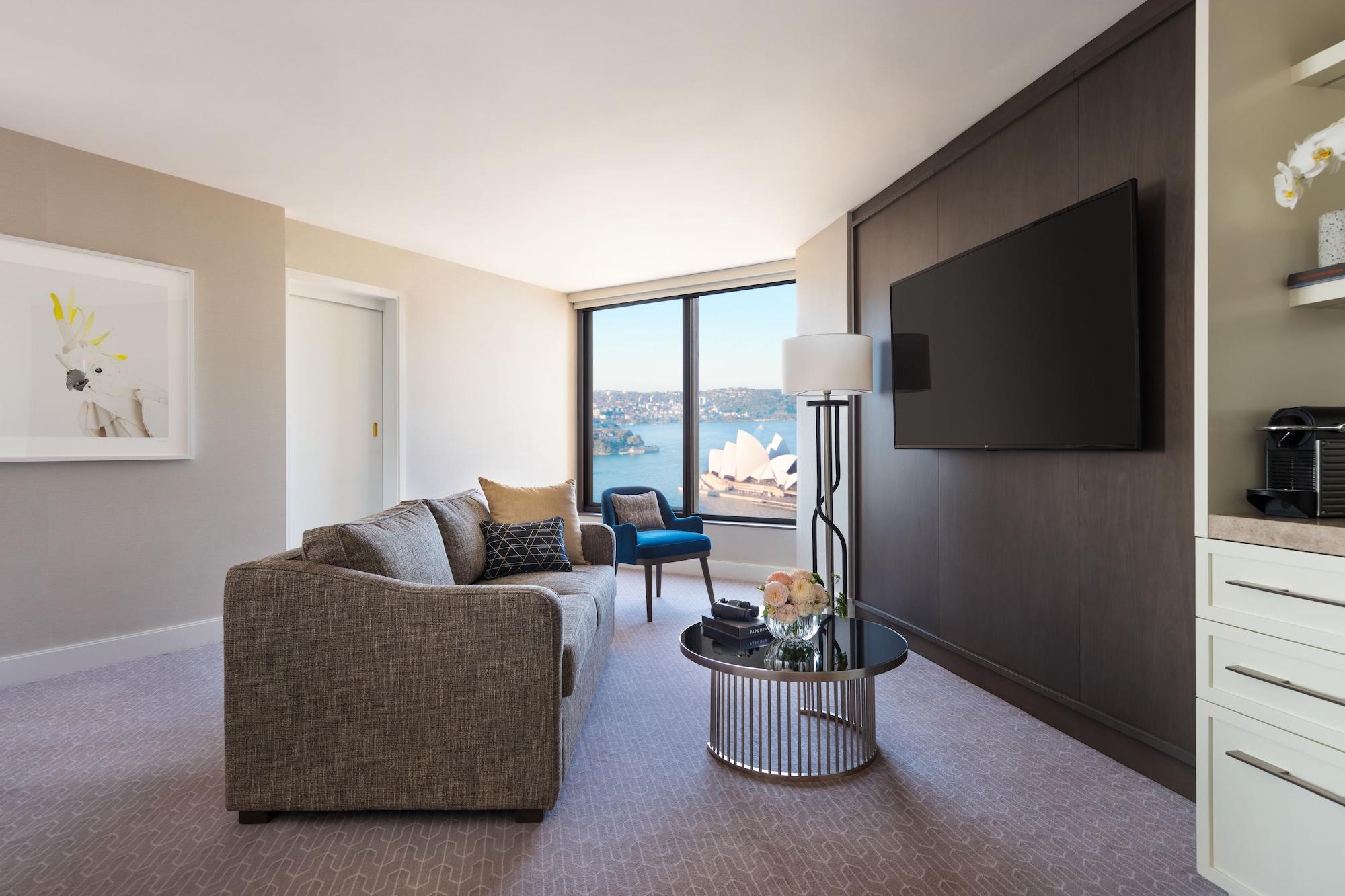 Four Seasons Hotel Sydney Exterior photo The photo shows a modern hotel room or suite. There is a comfortable gray sofa placed next to a round coffee table with a few decorative items. A tall lamp stands beside the sofa. On the opposite wall, there is a large flat-screen TV mounted, and lar