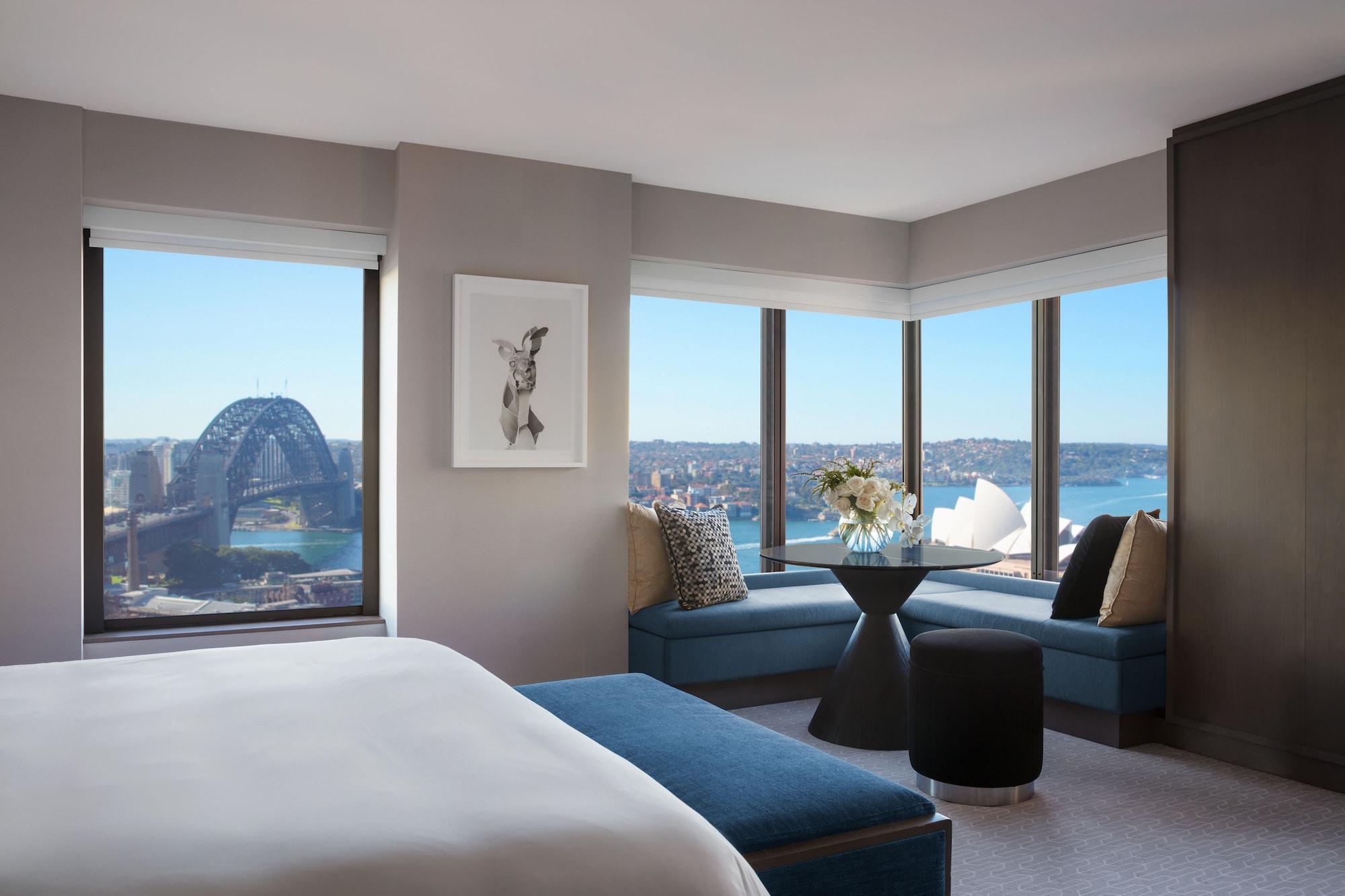 Four Seasons Hotel Sydney Exterior photo The photo shows a modern hotel room with large windows offering a panoramic view of a city skyline. In the foreground, there is a bed with white linens and a blue accent bench. A piece of artwork is displayed on the wall. In the corner, there is a ci