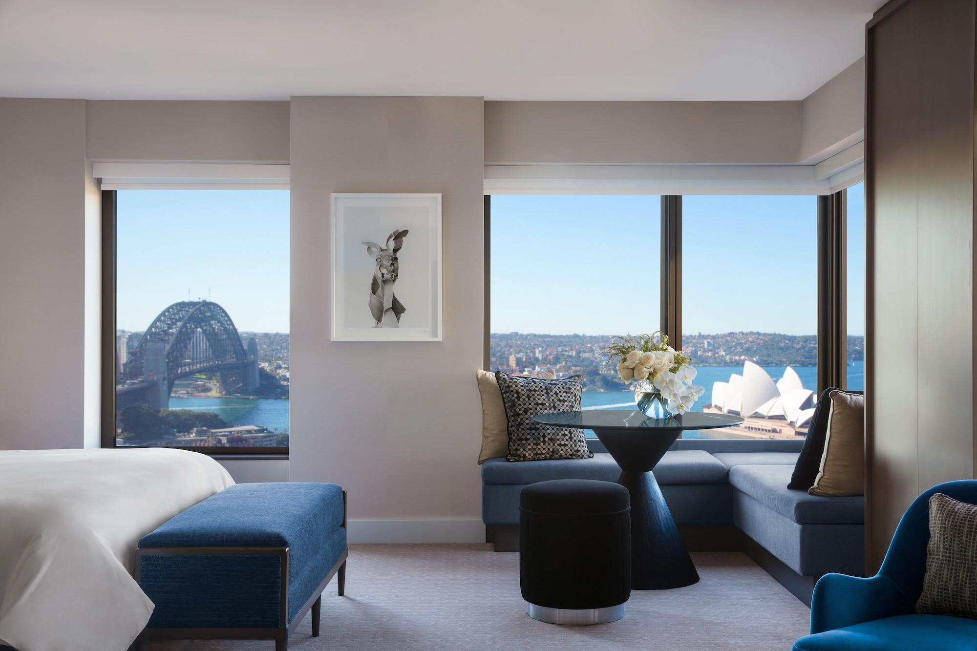 Four Seasons Hotel Sydney Exterior photo The photo shows a modern hotel room with large windows offering a view of the Sydney Opera House and the Sydney Harbour Bridge. The interior features a comfortable bed with a blue upholstered bench and decorative pillows. There's a round black table 