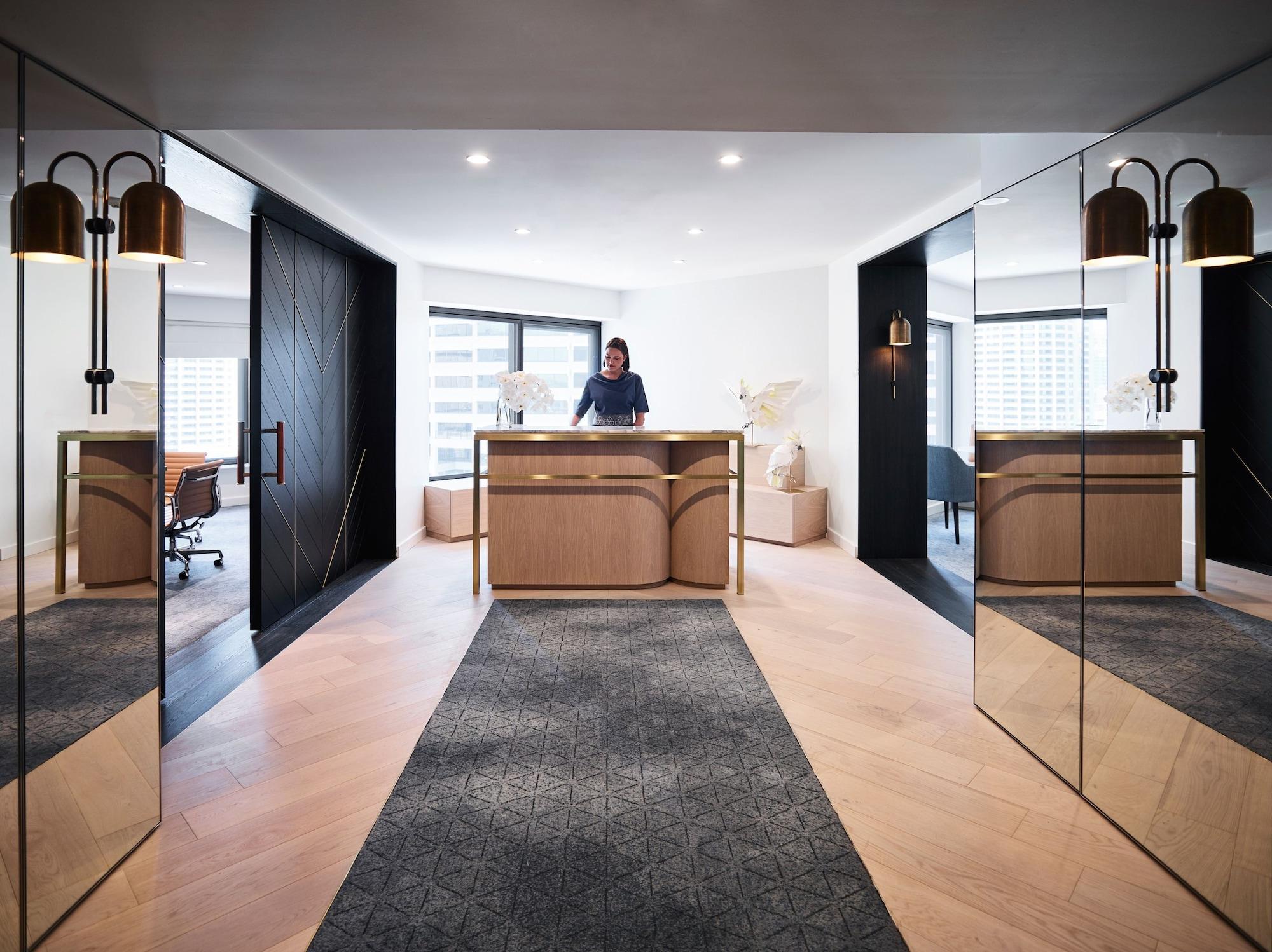 Four Seasons Hotel Sydney Exterior photo The photo shows a modern reception area, characterized by a sleek and stylish design. In the foreground, there is a wooden reception desk with a person standing behind it, likely greeting guests. The desk has a minimalist aesthetic, complemented by d