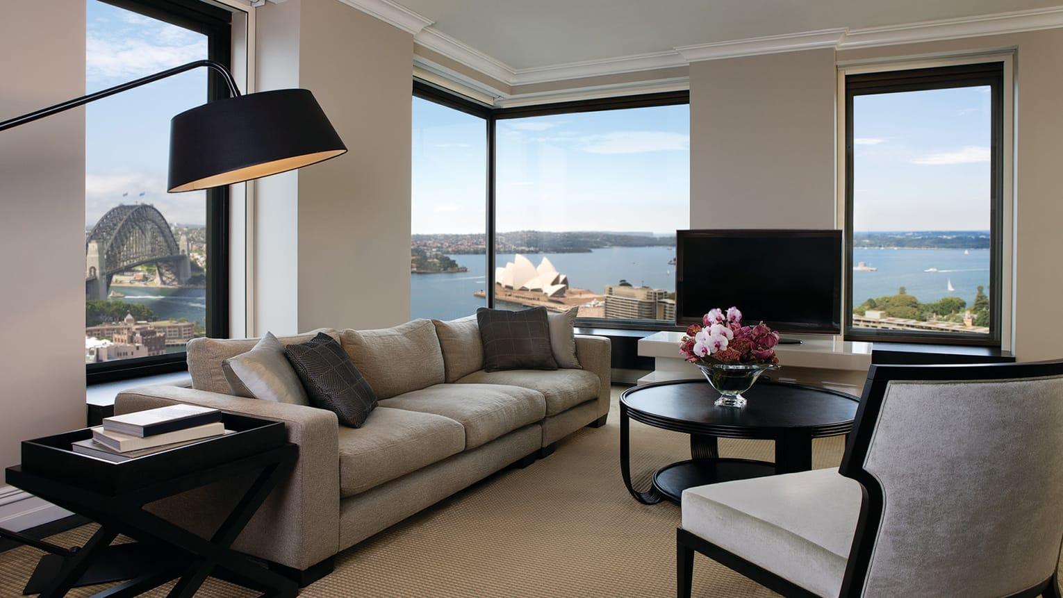 Four Seasons Hotel Sydney Exterior photo The photo shows a stylish and modern living room with large windows offering a view of a body of water and nearby buildings. The room features a comfortable, light-colored sofa and a round coffee table with a floral centerpiece. A flat-screen televis