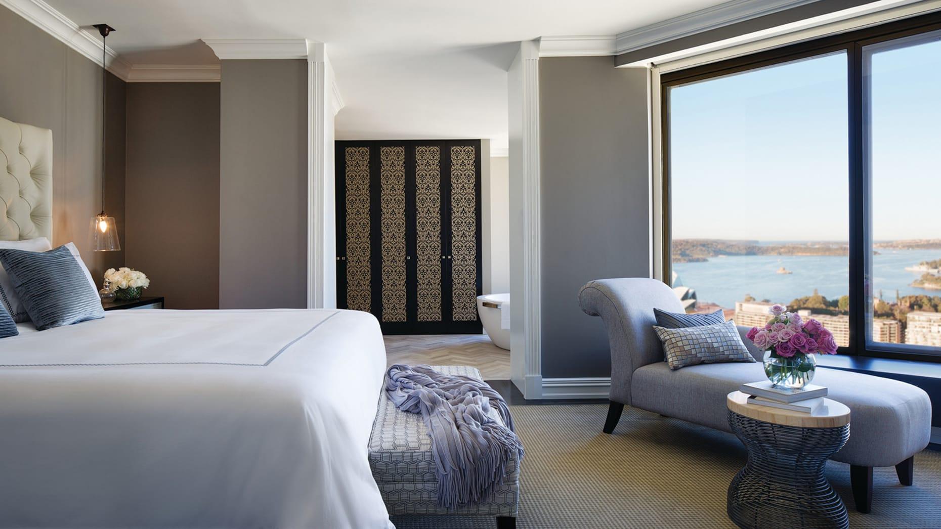 Four Seasons Hotel Sydney Exterior photo The photo shows a stylish hotel room featuring a large bed with crisp white linens. To the left, there is a comfortable seating area with a gray accent chair and a small round table topped with a vase of flowers. The room has a modern design, charact