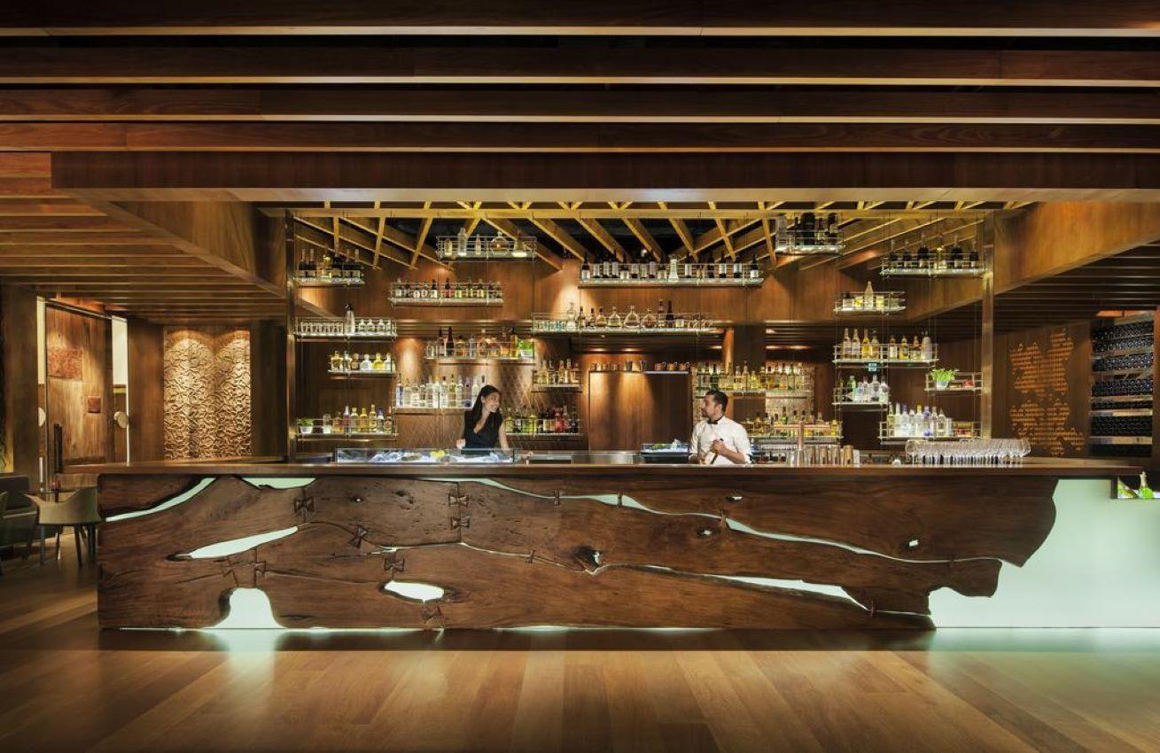 Four Seasons Hotel Sydney Restaurant photo The photo showcases a stylish bar area with a modern design. The bar counter is made of natural wood, featuring a unique, organic shape with visible grain patterns. Behind the bar, shelves hold an array of bottles, suggesting a well-stocked selection