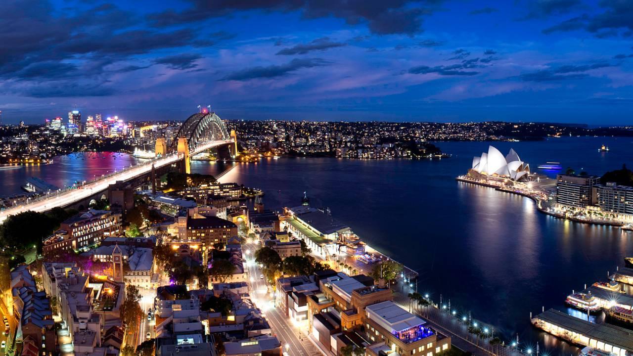 Four Seasons Hotel Sydney Exterior photo The photo depicts a stunning nighttime view of Sydney, Australia. In the foreground, you can see a bustling urban area with buildings and streets illuminated by lights. To the left, the iconic Sydney Harbour Bridge is visible, elegantly arching over 