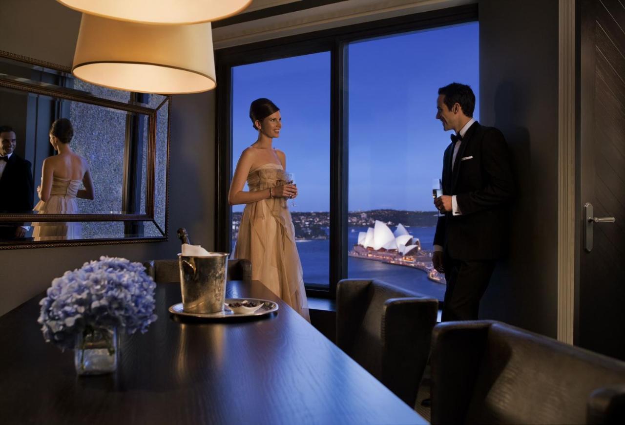 Four Seasons Hotel Sydney Exterior photo The photo depicts a stylish evening scene where a woman in an elegant dress and a man in a formal suit are engaged in conversation near a large window. They are in a modern interior space, with a table that has a bottle of champagne and a glass. The 