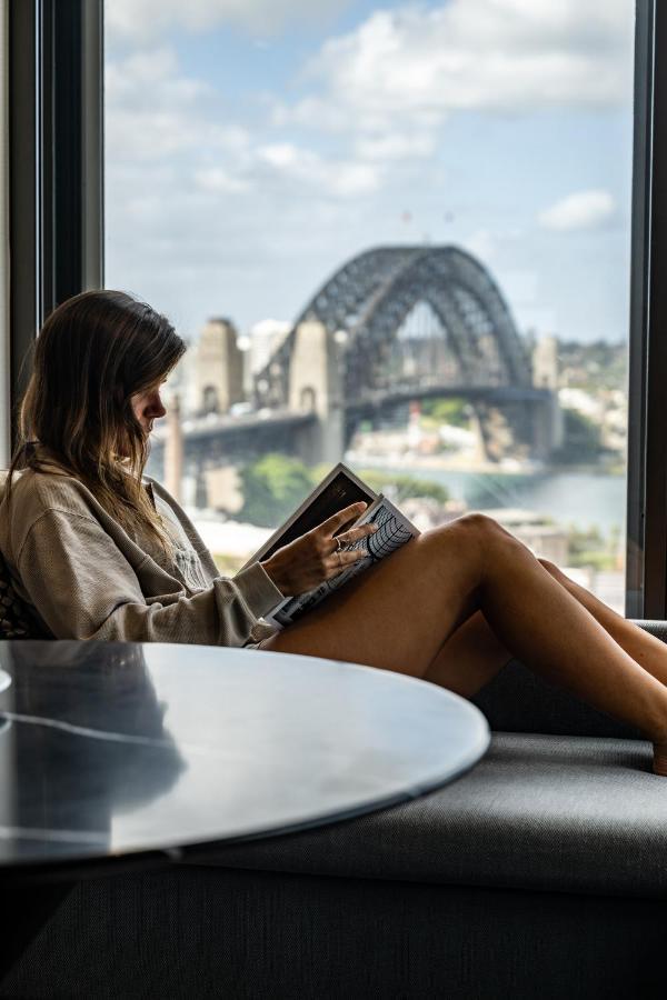 Four Seasons Hotel Sydney Exterior photo The photo depicts a woman sitting comfortably by a window, reading a book. She is wearing a casual oversized sweatshirt and is relaxed, with her legs crossed. The background features a view of a bridge, likely the Sydney Harbour Bridge, with a citysc