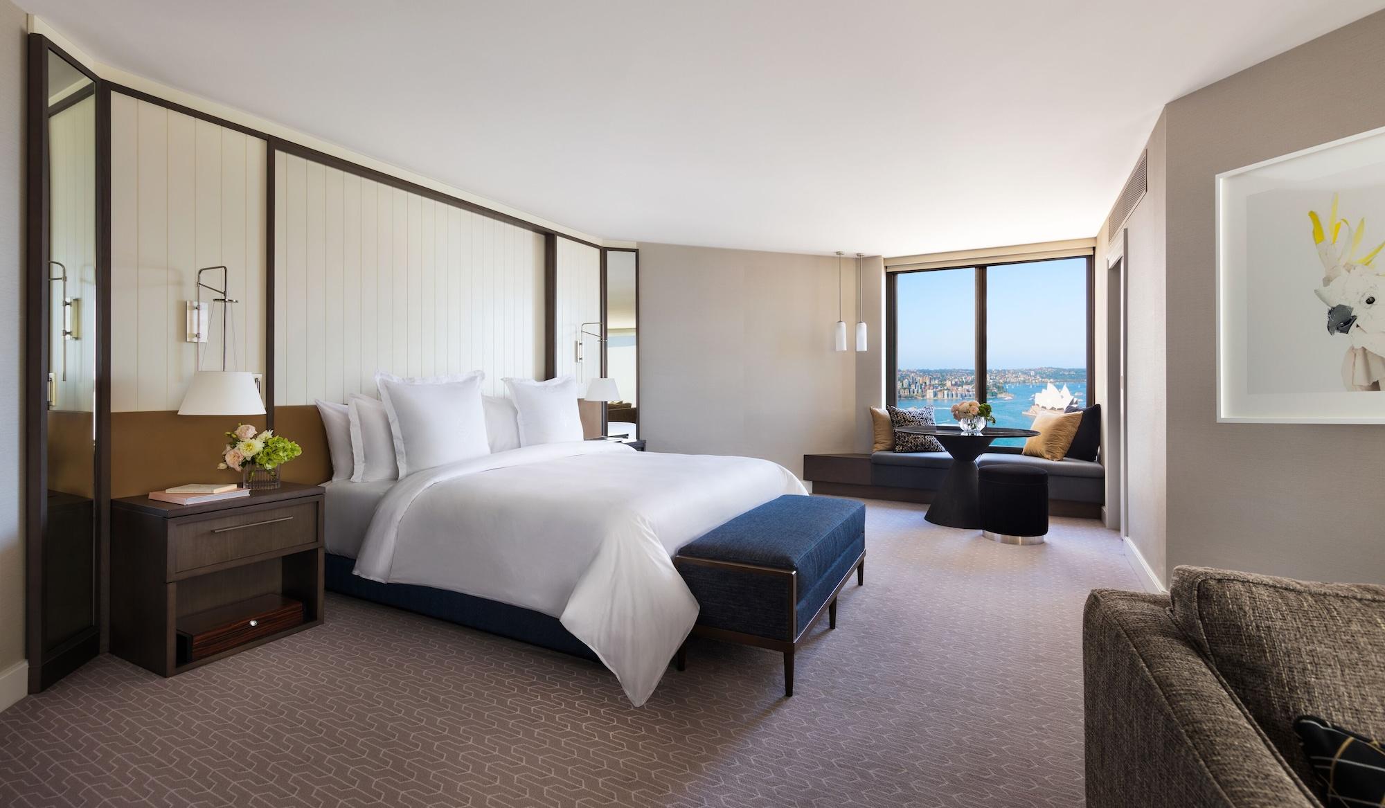 Four Seasons Hotel Sydney Exterior photo The photo shows a modern hotel room that features a spacious layout. In the center, there is a large bed with white linens, complemented by a blue upholstered bench at the foot of the bed. The walls have a light and neutral color scheme, creating a c