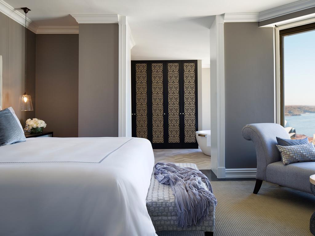 Four Seasons Hotel Sydney Exterior photo The photo shows a luxurious hotel room or suite. There is a large bed with white linens positioned towards the center. A gray armchair is nearby, accompanied by a small ottoman or stool. The walls have a soft gray tone, and there are elegant decorati