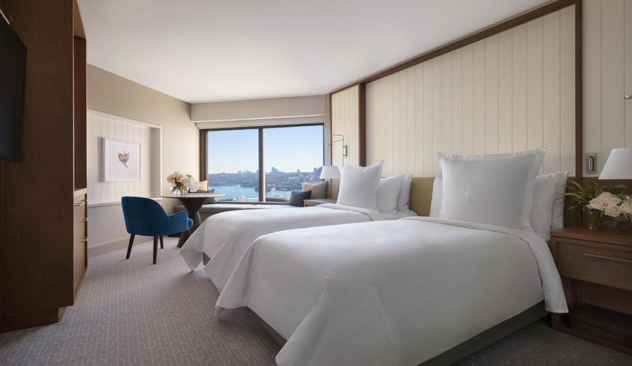 Four Seasons Hotel Sydney Exterior photo The photo shows a contemporary hotel room featuring two neatly made beds with white linens. There is a cozy blue chair positioned near a table, and a vase with flowers on the table adds a touch of decor. The large window offers a view of the outside,