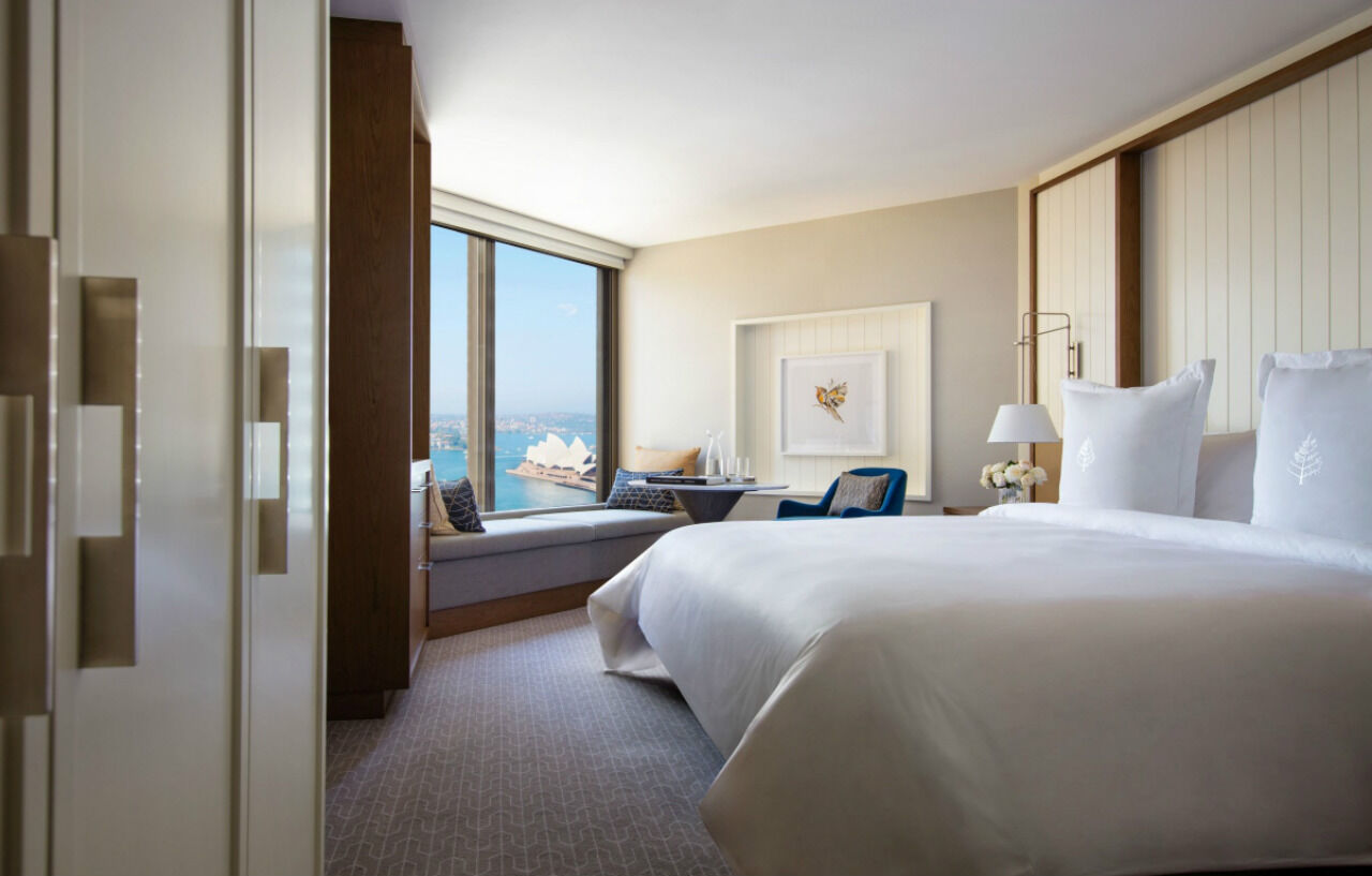 Four Seasons Hotel Sydney Exterior photo The photo shows a contemporary hotel room featuring a large window with a view. The room has a neatly made bed with white linens, a small seating area with a blue chair, and a cozy nook by the window. There’s a piece of artwork on the wall, and the d