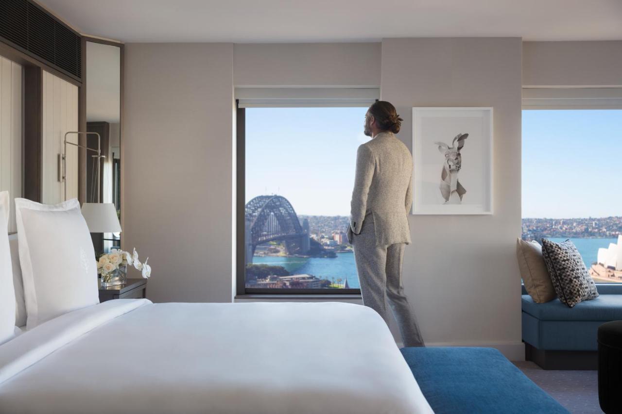 Four Seasons Hotel Sydney Exterior photo The photo features a modern hotel room with a large bed and a stylish decor. A person dressed in a formal outfit stands near a window, looking out. The view includes a scenic landscape, possibly of Sydney, showcasing the harbor and a landmark structu